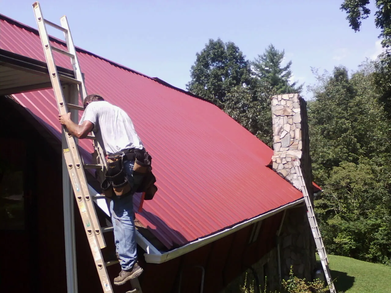 Roofer on a ladder beside a red metal roof and stone chimney — Soffit & Fascia Repair by Roofing Brothers in West Wareham